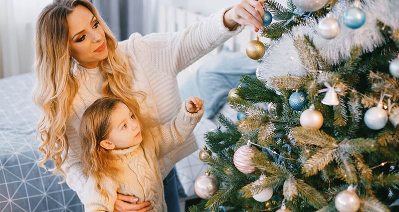 Mum with a daughter decorating a Christmas tree, a bed in the background