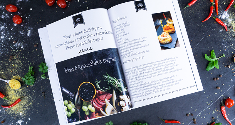 Otvorená fotokniha s receptami, vedľa nej ležia chilli papričky, cherry paradajky a korenie.