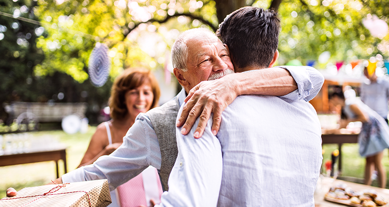 Papá abrazando su hijo adulto. Le regala una cosita durante una celebración familiar en un jardín.