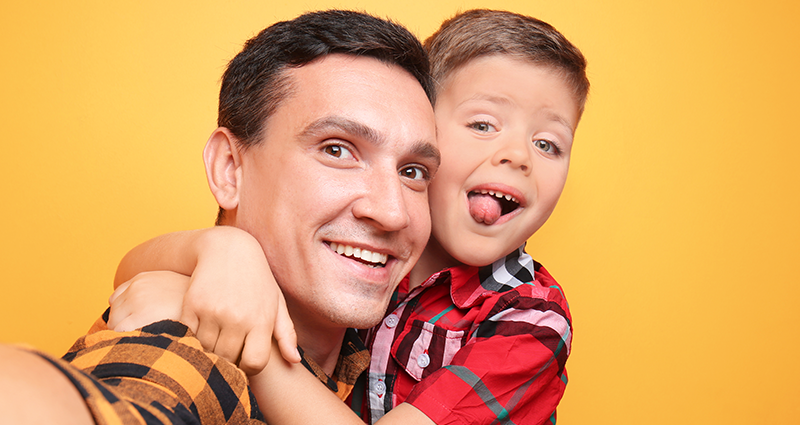 Un padre e suo figlio che indossano una camicia a quadretti e fanno un selfie. Sullo sfondo un muro arancione.