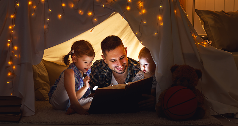 Papá leyendo a sus niños pequeños dentro de una tienda de campana hecha de sabana, luminada por las luces.