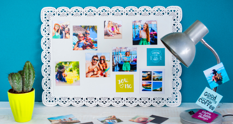 Photo magnets on a metallic white board, a part of them lying on the desk; a cactus in a green flowerpot on the left, a lamp with pinned magnets on the right.