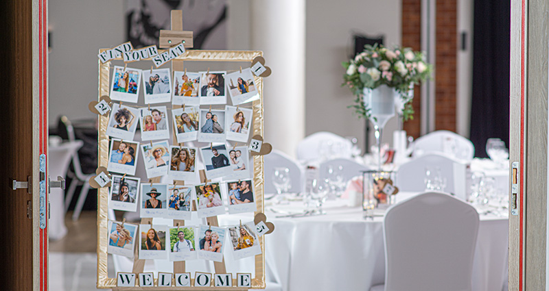 Foto di una cornice handmade con le foto degli ospiti del matrimonio con vicino i numeri dei tavoli. Nella parte superiore è visibile la scritta "Find your seat", nella parte inferiore la scritta "Welcome"; sullo sfondo si vede un tavolo e la sala del ristorante.