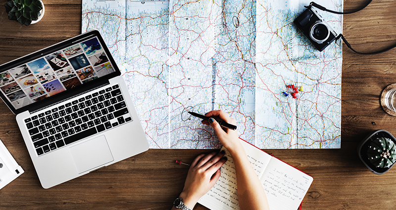 Photo of a person getting ready to his/her journey: world map in the center, laptop, camera and notebook on a desk