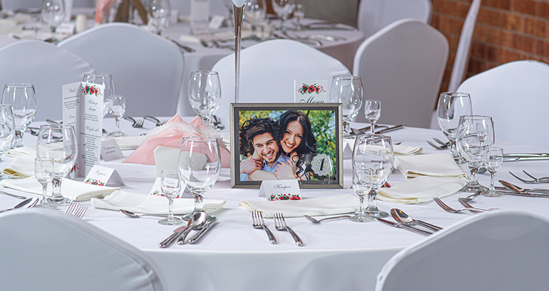 Foto de los novios en un marco negro plateado, con el número 1, en el centro de la mesa decorada en blanco, alrededor de la vajilla y viñetas.