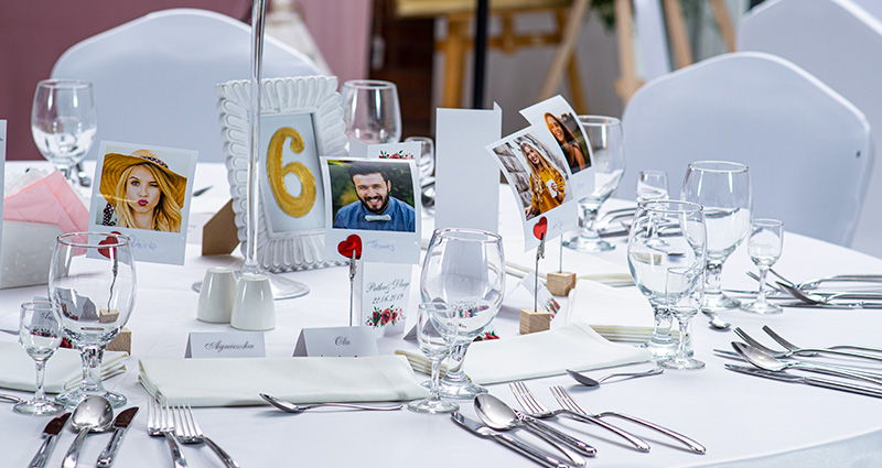 Dei segnaposto con le foto degli invitati al matrimonio, stampate come foto retrò su cui sono stati aggiunti anche i cognomi. Le decorazioni sono state sistemate su un tavolo decorato di colore bianco, con sopra i bicchieri e le posate.