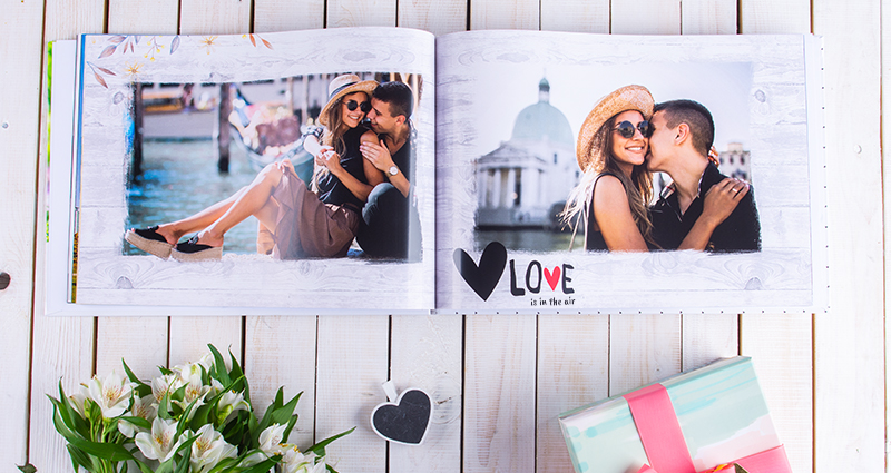 Fotos de una pareja enamorada impresas en un fotoálbum, debajo de una de las fotos hay un texto: Love is in the air. El fotolibro sobre un fondo blanco y de madera, debajo del fotolibro hay decoraciones de flores y un envelto regalo multicolor.