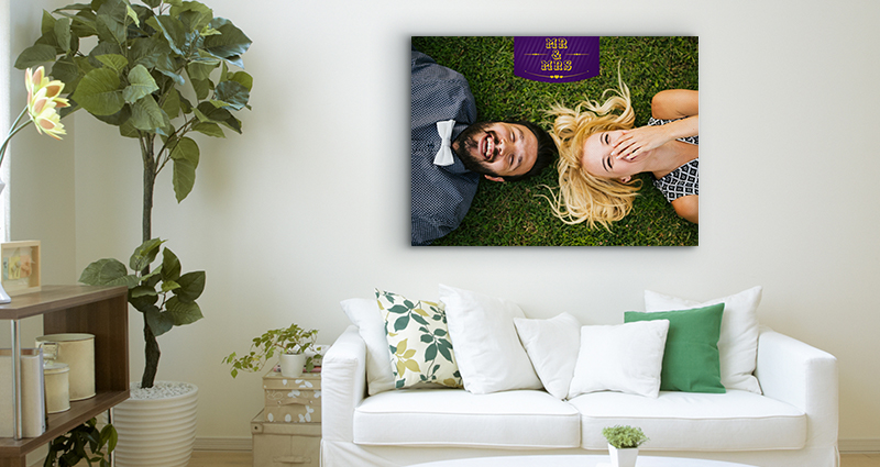 Picture of a bright living room with green cushions on a white couch. A big plant in a flowerpot nearby and a huge photo canvas with a photo of a couple, laying on the grass, hanging over the couch.