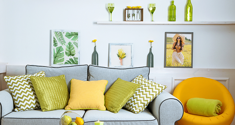 Picture of a grey couch with green and yellow cushions in various patterns. A yellow armchair next to the couch. A coffee table with fruits on it in front of the couch. 3 spring pictures and various decorations behind the couch.