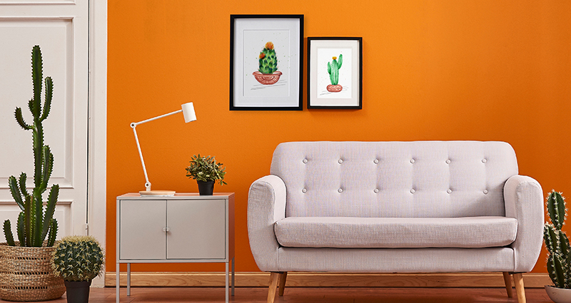 Picture of a living room with a close-up on a bright couch and an end table. Cactuses on both sides of the couch. Orange wall in the back with two spring pictures in black frames.