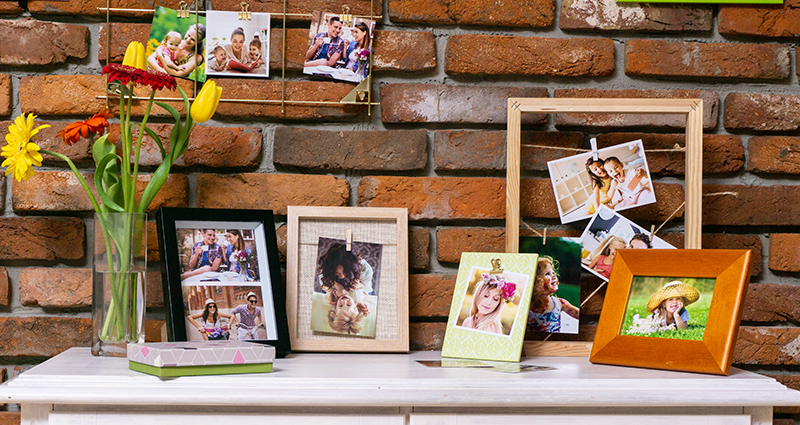 Foto von einer weißen Kommode, eine Backsteinwand im Hintergrund. Auf der Kommode Frühlingsfotos in bunten Rahmen,  daneben eine Vase mit gelben und orangen Blumen. An der Wand hängen Insta Fotos in Rahmen.
