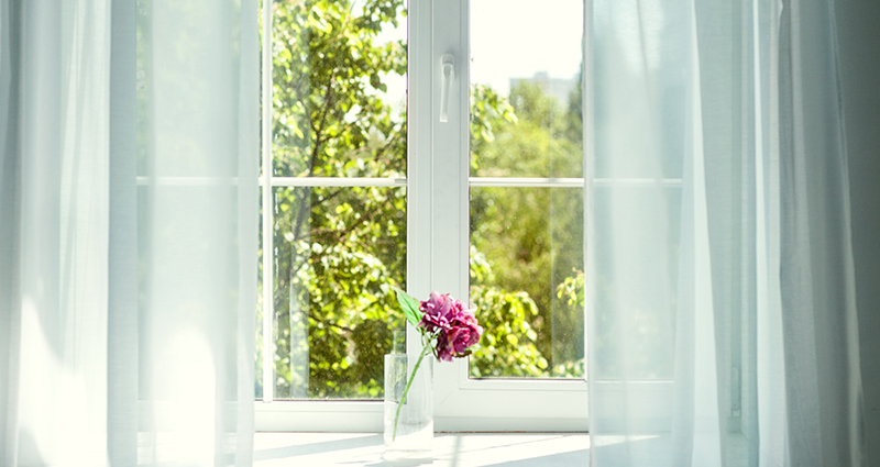 Een foto van een raam met lichte gordijnen. Buiten het raam staat een boom en op de vensterbank bevindt zich een roze bloem.