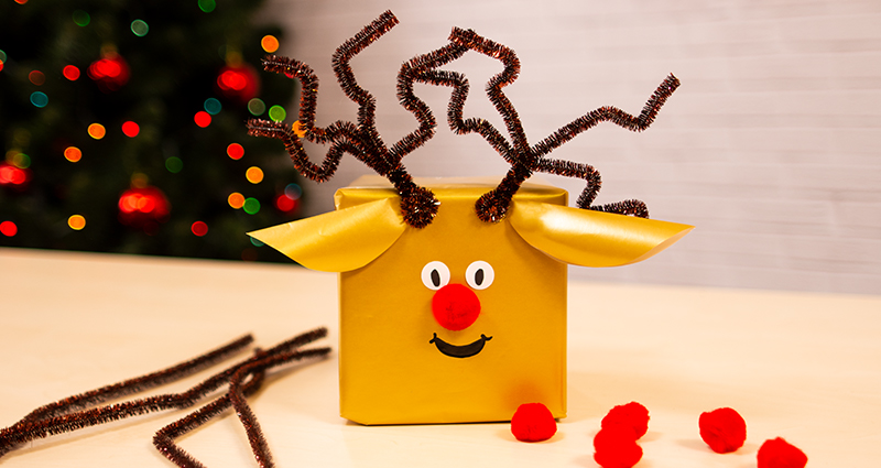 Una caja navideña de regalo en forma de reno. Al lado de la caja, limpiapipas para manuales de color marrón y pompones rojos, un árbol navideño en el fondo.