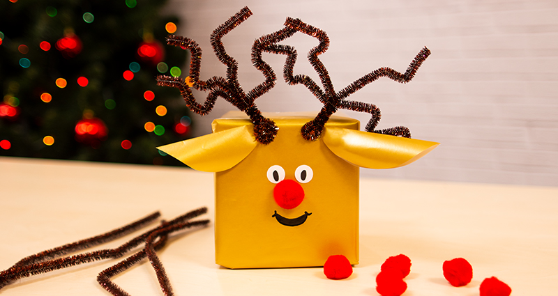  Cadeaudoosje in de vorm van een rendier gepresenteerd op een heldere tafel. Naast zijn er bruine pijpenragers en rode pompons te zien. Op de achtergrond - een kerstboom. 