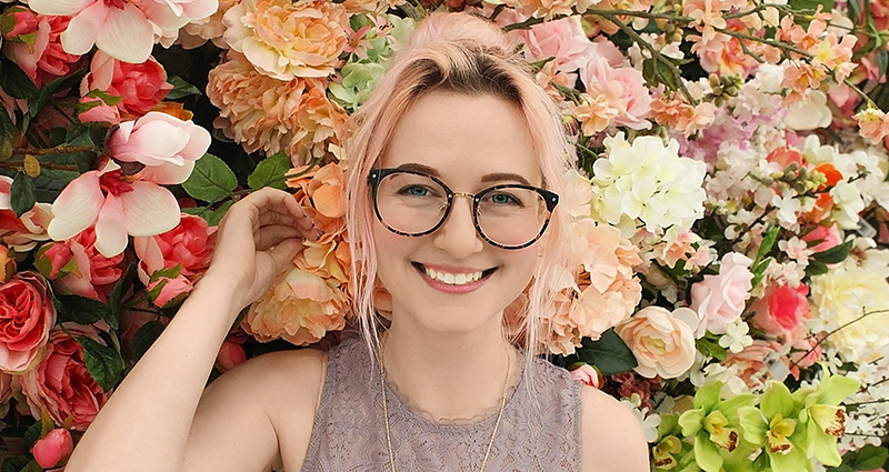 Mujer sonriendo con las gafas y un arbusto de flores en el fondo.