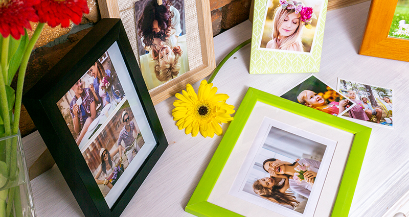 Spring photo prints in colourful frames on a white dresser. Red gerberas in a vase on the left side and a yellow gerbera laying in between the frames.
