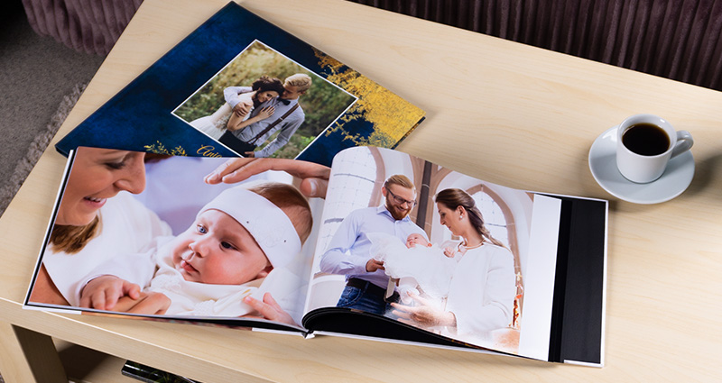 Dos Starbooks puestos en una mesa clara – uno abierto con fotos de bautismo y otro cerrado con fotos de boda; una taza de café y flores en un jarrón cerca de los álbumes