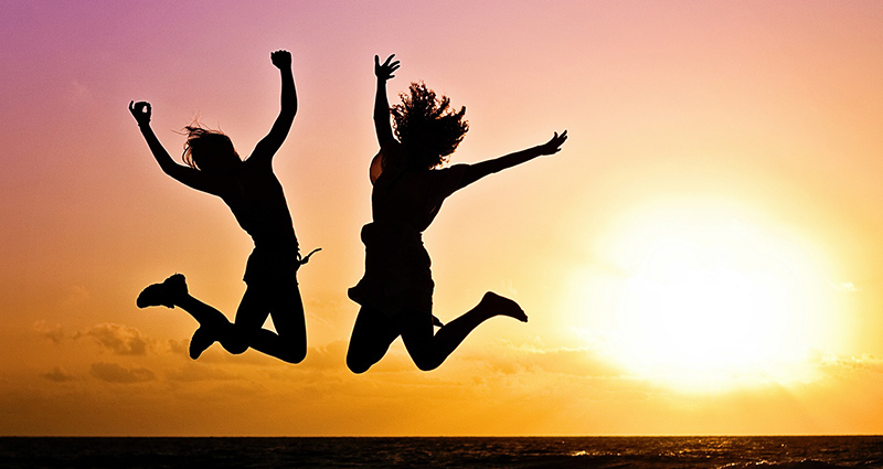 Dos chicas saltando en la playa sobre el horizonte, el mar y la puesta de sol en el cielo amarillo-rosado en el fondo.