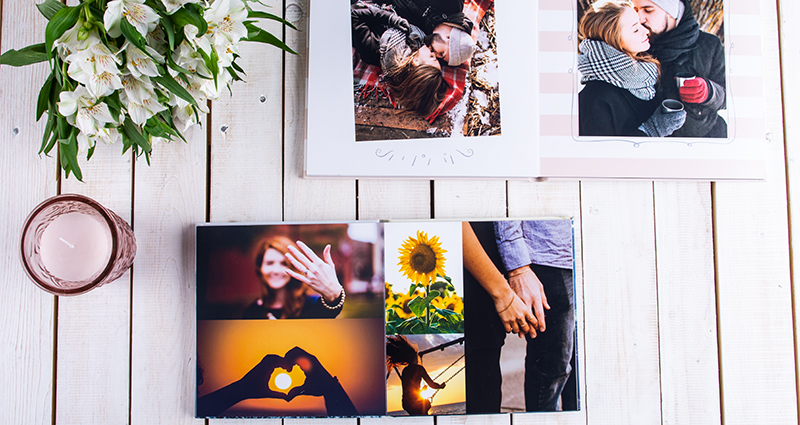 Zwei aufgeschlagene Echtfotobücher mit Fotos eines verliebten Paares, links ein Strauß weißer Blumen und eine Kerze.