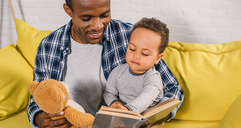 Un hijo en los brazos de papá que está leyendo un libro. Sentados juntos en el sofá amarillo.