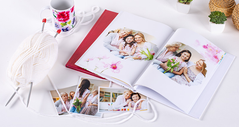 Un livre photo et des tirages avec des photos de grand-mère et ses petits enfants, à côté un mug en fleurs et un fil en couleur claire avec des aiguilles à tricoter.