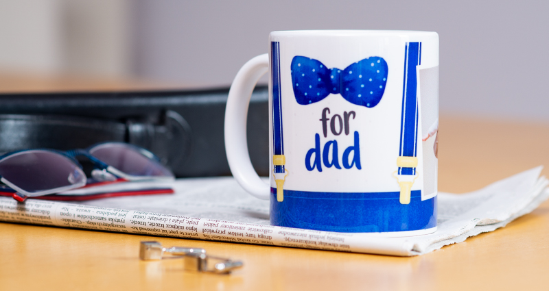 Una fototazza colorata, progettata utilizzando il modello per la Festa del Papà, caratterizzata da un paio di bretelle e un papillon a pois. La tazza è appoggiata su un giornale, con accanto un paio di occhiali, una borsa e dei gemelli da polso.