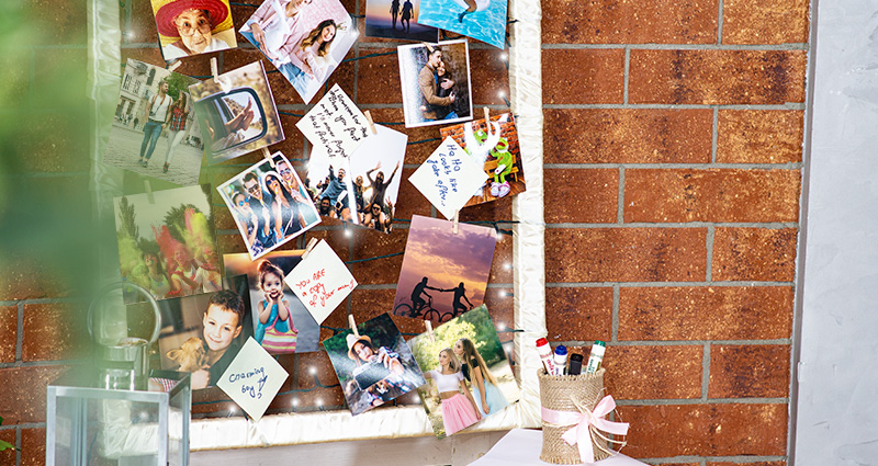 Fotografías que convocan la historia de amor de los novios en un marco de madera decorado con cinta de satín y luces LED. Al lado una lata con marcadores coloridos adentro. En el fondo una pared de ladrillo rojo. 
