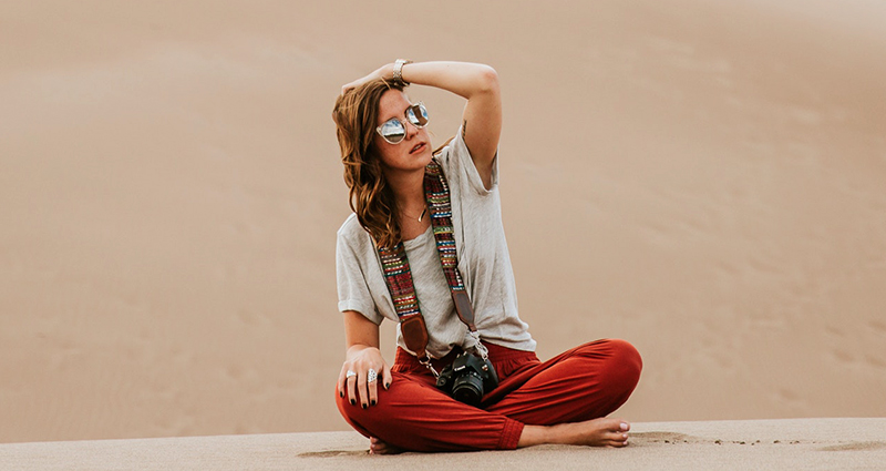 Woman sitting cross-legged and fixing her hair. There is a camera with a thick handmade strap on her neck.