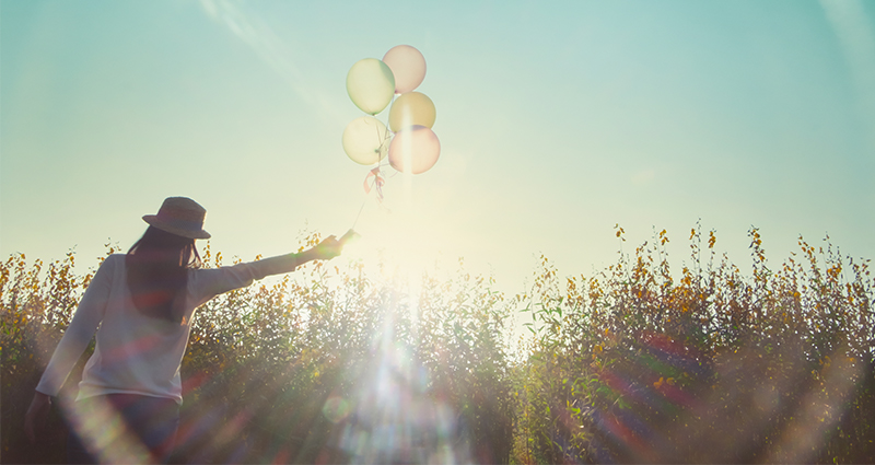 Een vrouw in een hoed op een weide met ballonnen in haar handen, foto genomen onder de zon.