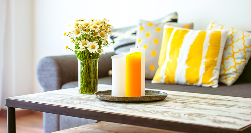 Geelgrijze kussens met kleurrijke patronen op een grijze bank, een vaas met madeliefjes en gele en witte kaarsen.