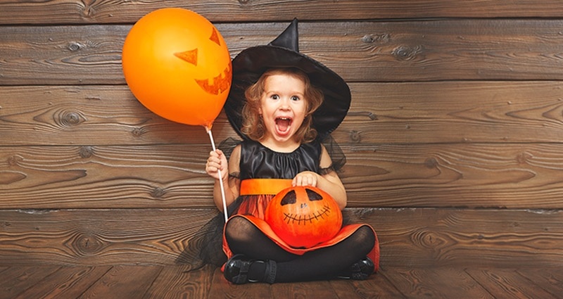 A girl dressed as a witch, holding a balloon and a pumpkin.
