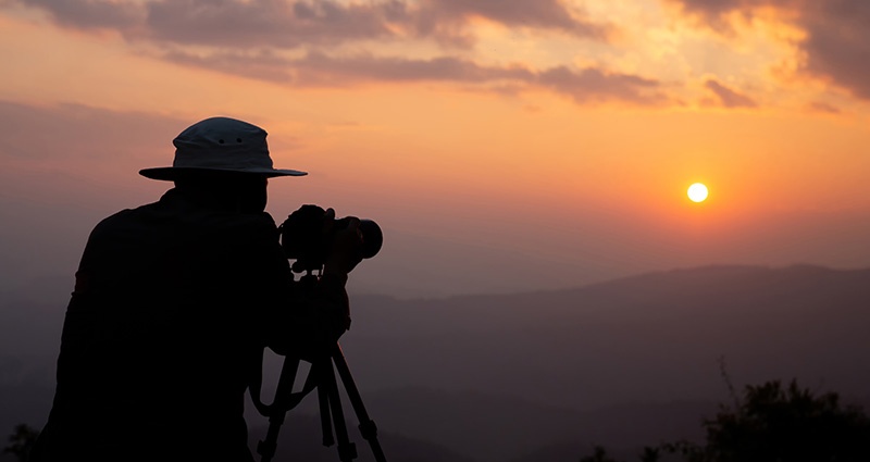 A man wearing a hat, taking a photo at sunset