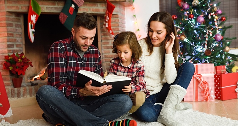 Family of three sitting in front of the fireplace with Christmas socks hanging above. A Christmas tree and gifts on the right in the background.