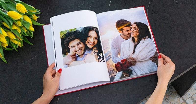 Focus on a woman’s hands holding an open photo book with photos of people in love; two mugs, a gift wrapped with a red bow and yellow tulips in a bucket next to it.