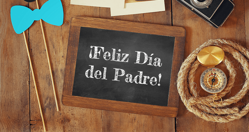 La tabla de madera con inscripción  "Feliz Día del Padre", rodeada de una cámara, una cuerda, unos revelados retro, una brújula, una pajarita y unos bigotes en palitos para selfie, unos accesorios sobre escritorio de madera.