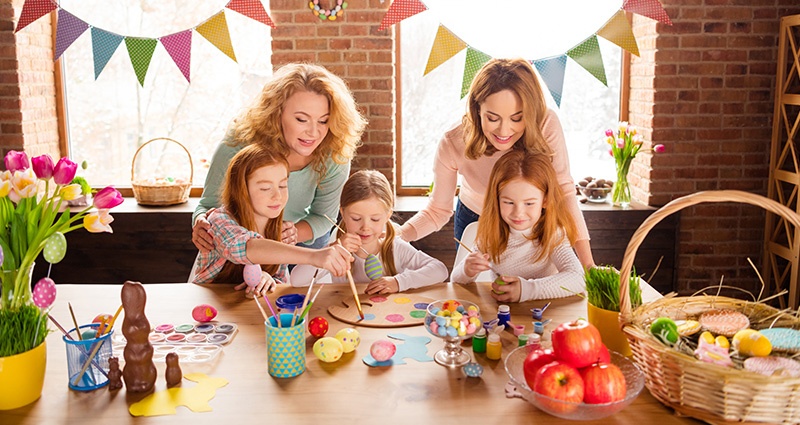 Trys mergaitės ir dvi moterys prie stalo, pilno kiaušinių, šokolado saldainių, dažų ir tulpių.